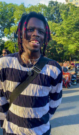 Pink Gibbs stands in Washington Square Park wearing a blue and white striped sweater and silver jewelry with facial piercings and pink in their dreadlocks.