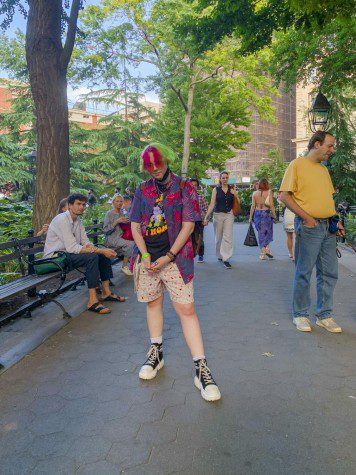 August Apostolakis-Beatty who has pink and green hair, stands in Washington Square Park wearing patterned shorts, a graphic t-shirt and an unbuttoned Hawaiian shirt.