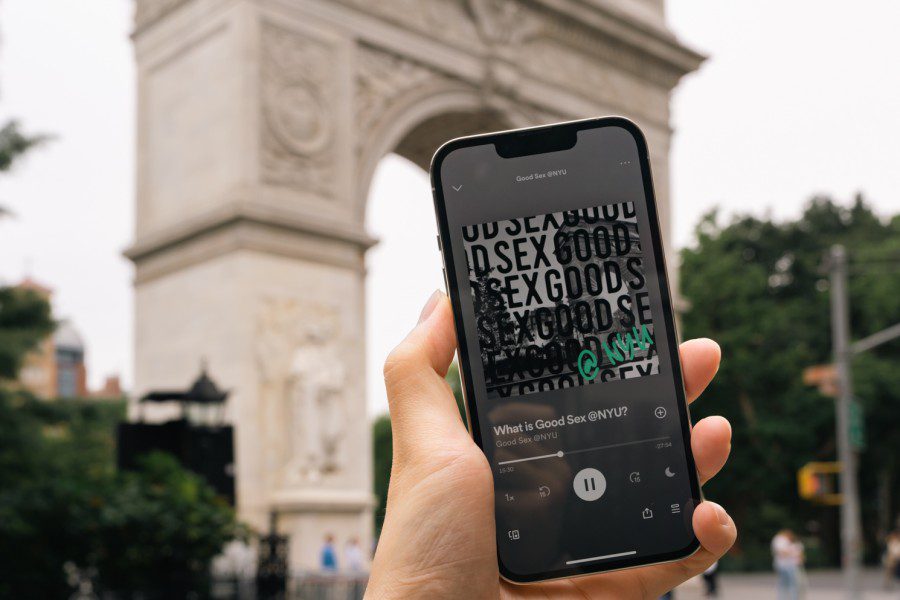 A hand holding a smartphone with the podcast “What is Good Sex @ NYU” playing on the screen, with the Washington Square Arch in the background.