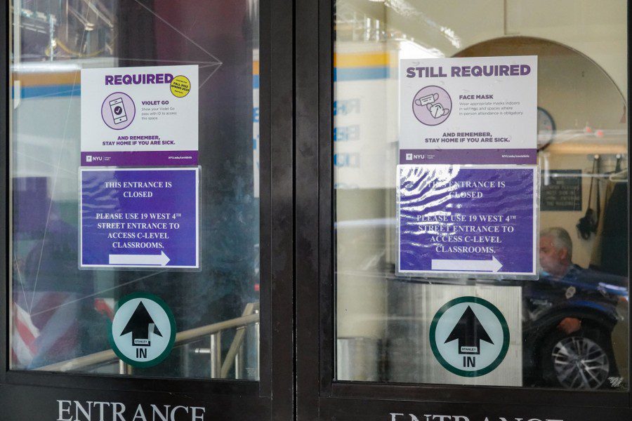 The entrance to a building with two signs showing arrows and the text IN and two identical notices of “THIS ENTRANCE IS CLOSED PLEASE USE 19 WEST 4TH STREET ENTRANCE TO ACCESS C-LEVEL CLASSROOMS” sticked on two glass doors.