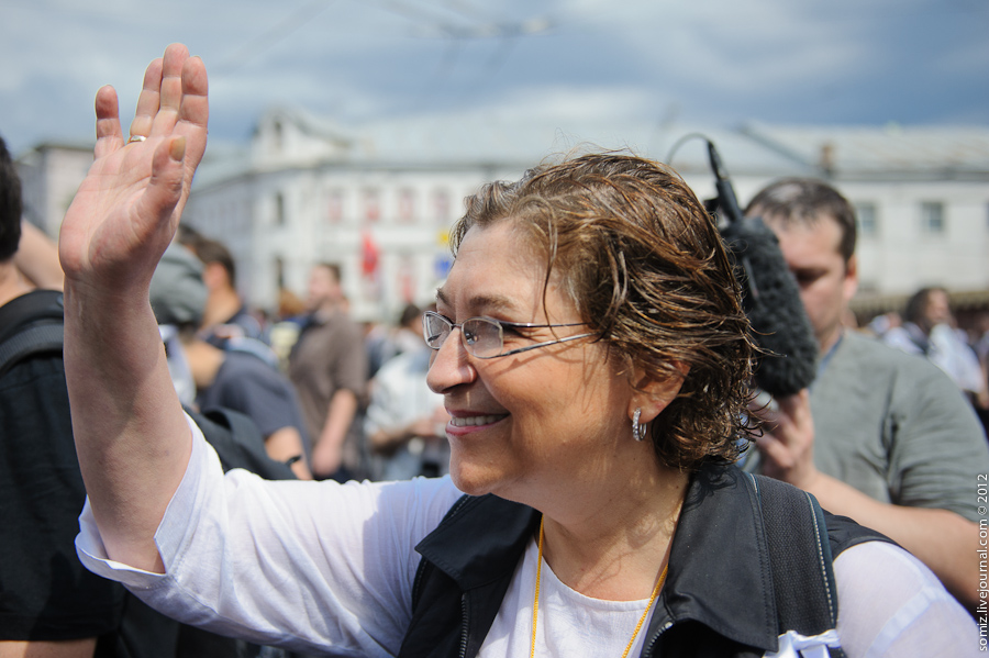 Yevgenia Albats dressed in a white t-shirt and a black vest, waving to a crowd.