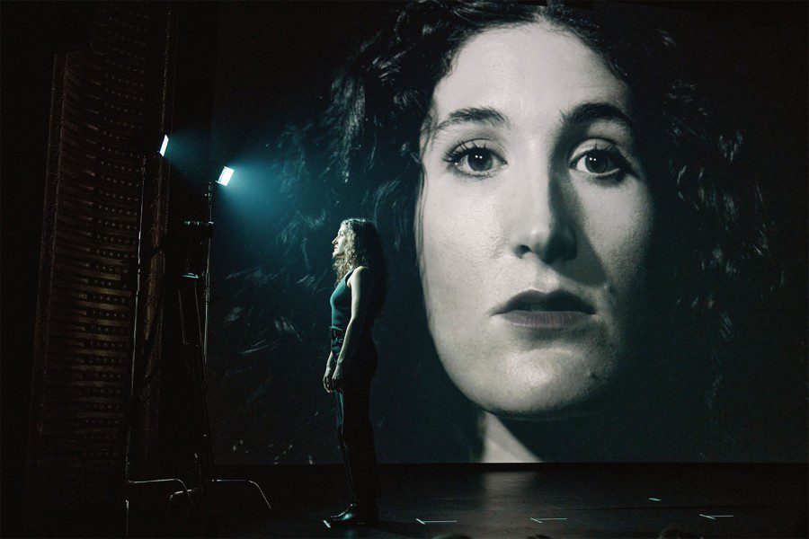 Kate Berlant dressed in all black stands on stage facing two bright stage lights with a projection of her face enlarged in the background.