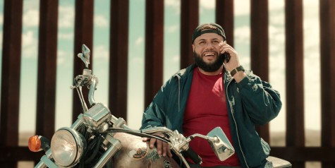 A bearded man in a red shirt and teal jacket takes a call while sitting on a parked motorcycle.
