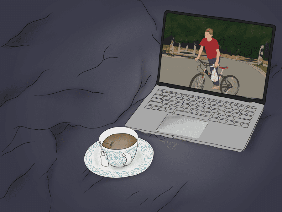 A laptop and a cup of tea lay against a dark gray blanket. On the laptop screen is a picture of a boy riding a bike in a forest.