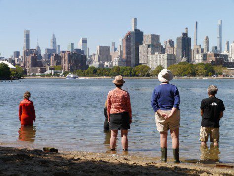Five people standing in shallow water in the East River, with midtown Manhattan skyline in the background. From the left: a person with short blonde hair wearing a red coat; a person wearing a black coat; a person with a hat, a red shirt with white dots and a black skirt; a person wearing white hat, blue jersey and beige shorts; a person with black t-shirt and beige shorts.
