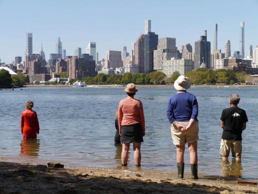 Five+people+standing+in+shallow+water+in+the+East+River%2C+with+midtown+Manhattan+skyline+in+the+background.+From+the+left%3A+a+person+with+short+blonde+hair+wearing+a+red+coat%3B+a+person+wearing+a+black+coat%3B+a+person+with+a+hat%2C+a+red+shirt+with+white+dots+and+a+black+skirt%3B+a+person+wearing+white+hat%2C+blue+jersey+and+beige+shorts%3B+a+person+with+black+t-shirt+and+beige+shorts.