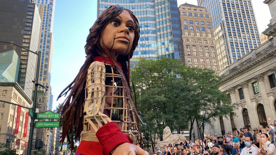 A large wooden puppet named Little Amal with brown skin, long brown hair and clothes with red accents stands in front of the New York Public Library with skyscrapers and street signs in the background. There is a male operating the puppet from inside and a crowd looking at and taking photos of the puppet.