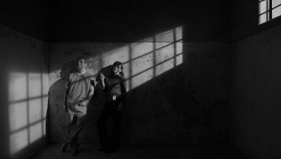 A monochromatic photograph of a prison cell. Light comes through the iron bar on the top right. A male prisoner and a female officer smoking a cigarette stand together on the left.