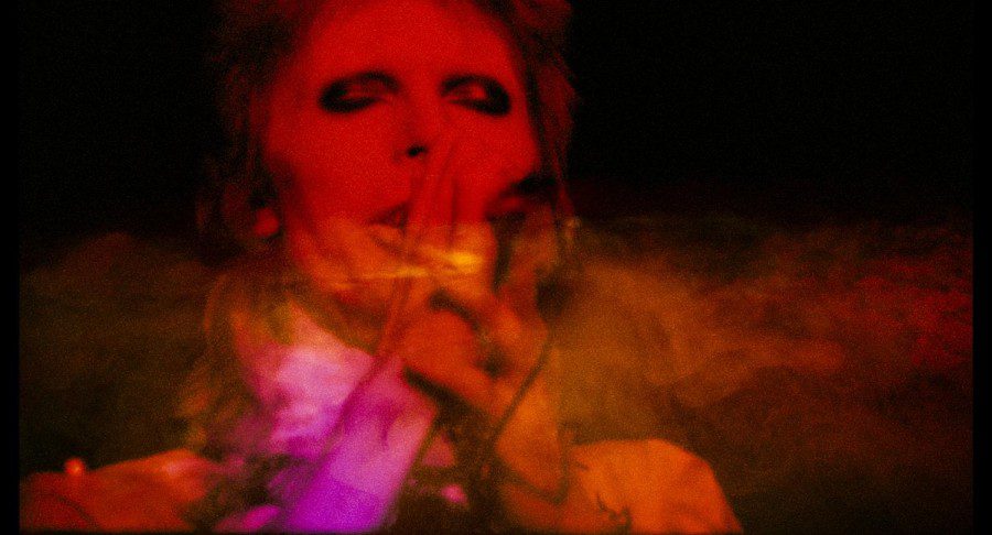 David+Bowie+with+dark+eyeshadow+and+long+hair+holds+a+microphone+to+his+mouth+while+his+eyes+are+closed+and+is+surrounded+by+neon-colored+fumes.