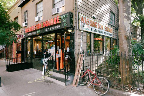 A facade of a restaurant at a street corner with a sign of neon lights, text “Washington Square Diner,” and tree shades next to the building. A perpendicular sign reads “Washington Square.”