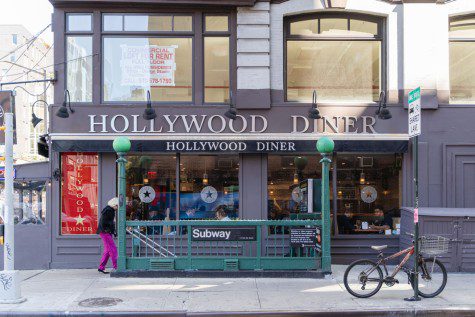 :The facade of a restaurant behind a subway station entrance with the text “HOLLYWOOD DINER” hanging on the wall. Smaller signage beneath it also reads “HOLLYWOOD DINER.”