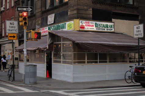 The facade of a restaurant at a street corner with outdoor dining spaces and text “Joe Junior Restaurant” on the light sign. A perpendicular sign reads “Joe Junior Restaurant.”