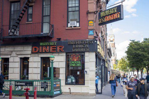 The facade of a restaurant at a street corner behind a subway station entrance with text “WAVERLY DINER” on the light sign in red and “BREAKFAST LUNCH DINNER” in brown.