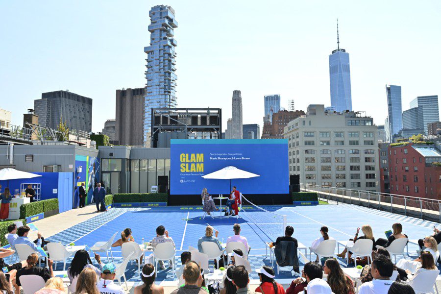 Maria Sharapova and Laura Brown sit under a parasol on a tennis court on a rooftop with a large audience in front of them. The downtown Manhattan skyline is behind them.