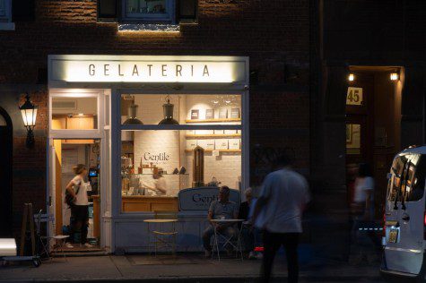 he facade of a warmly lit shop with white walls and a sign with text “Gelateria.” The storefront window reads “Gentile” and “GELATIERI DAL 1880.” There is one person standing in the shop and two people sitting outside.