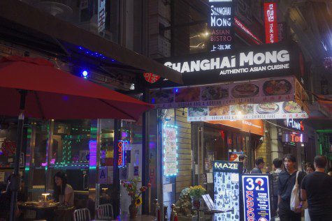 The entrance of a restaurant at nighttime with outdoor seating and people passing by. The entrance is surrounded with neon light signs with text “SHANGHAI MONG” and “Authentic Asian Cuisine.”