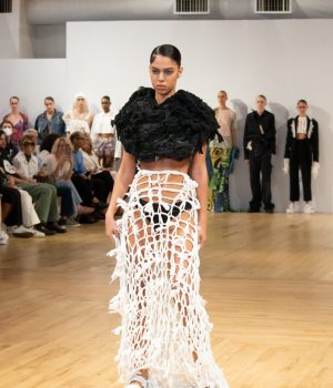 A model wearing a white open-tied skirt, white platform sandals and a knotted black cropped top pauses at the end of the runway.