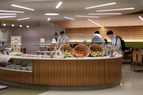 Centered in the photo are two students at a salad bar with dining tables to the right.