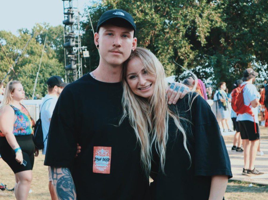 A man with tattoos on his arm hugs a woman with blonde hair-leaning on the man’s left shoulder while they stand on the ground of a music festival.