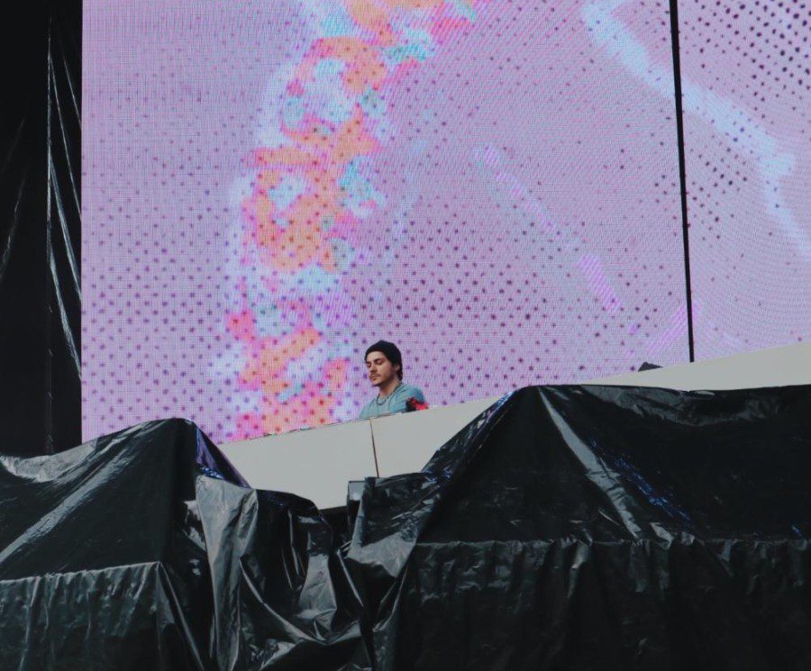 A man with a green sweatshirt and a black beanie stands on stage behind his equipment and in front of large LED screens.