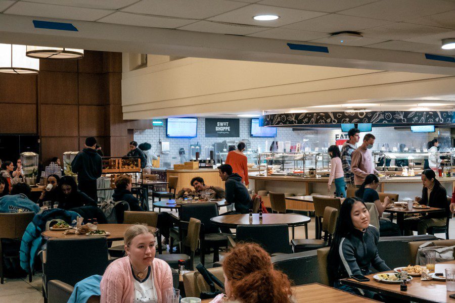 There is an eating area with students eating and chatting at circular tables in the foreground and a buffet in the background.