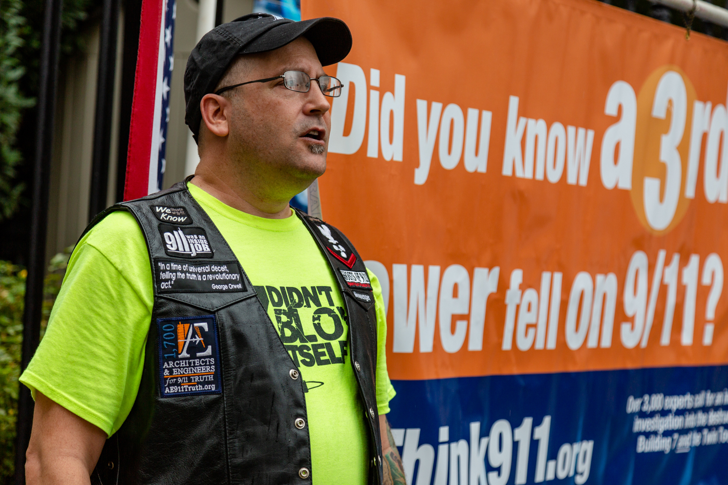A member of the conspiracy group Architects and Engineers for 9/11 Truth, dressed in a neon-green t-shirt and a black leather vest, stands next to a poster that says “Do you know a third tower fell on 9/11?”
