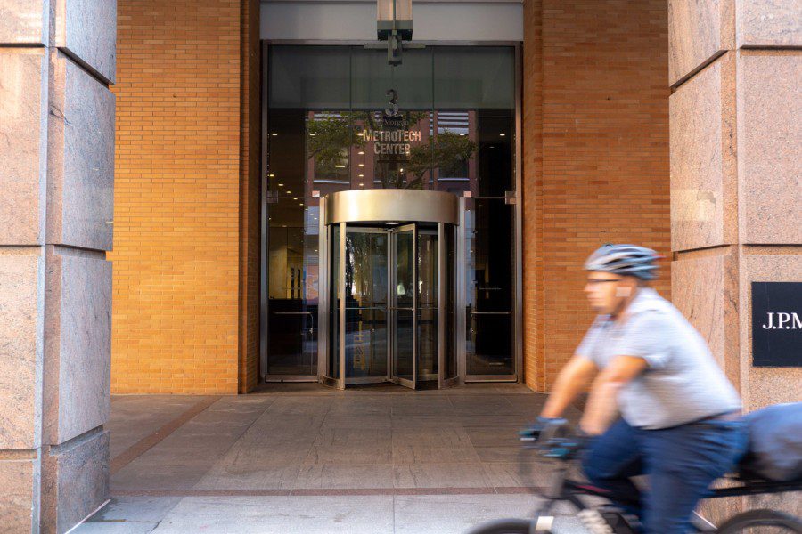 The entrance to a building with a revolving door and text “3 MetroTech Center” hanging on the glass windows, while a person riding a bicycle passes by.