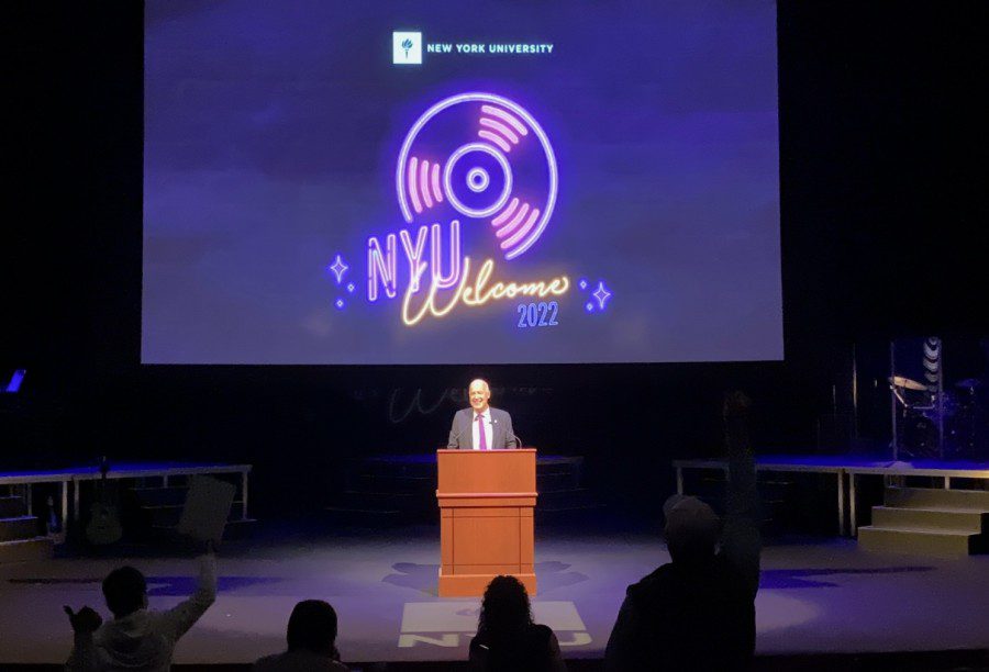 NYU President Andrew Hamilton stands on stage behind a podium with a screen with the text “NYU Welcome 2022” and a logo of a music disc displayed behind him.