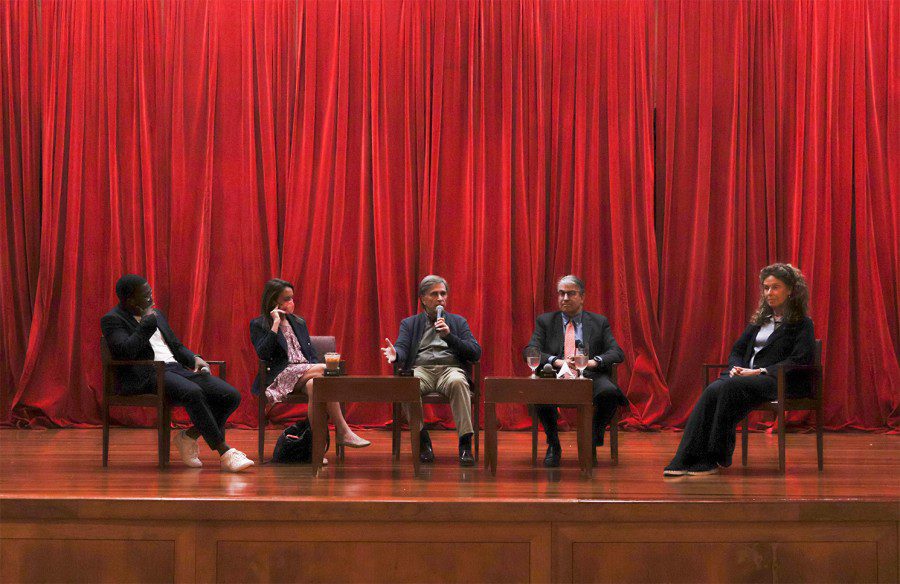 Three men and two women sit on a stage with red drapes in the background while the man in the middle speaks using a microphone.