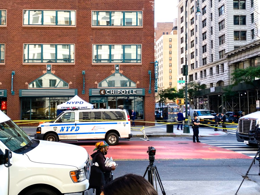 Police officers block off a New York City street corner with yellow caution tape in front of a Chipotle and apartment building facade.