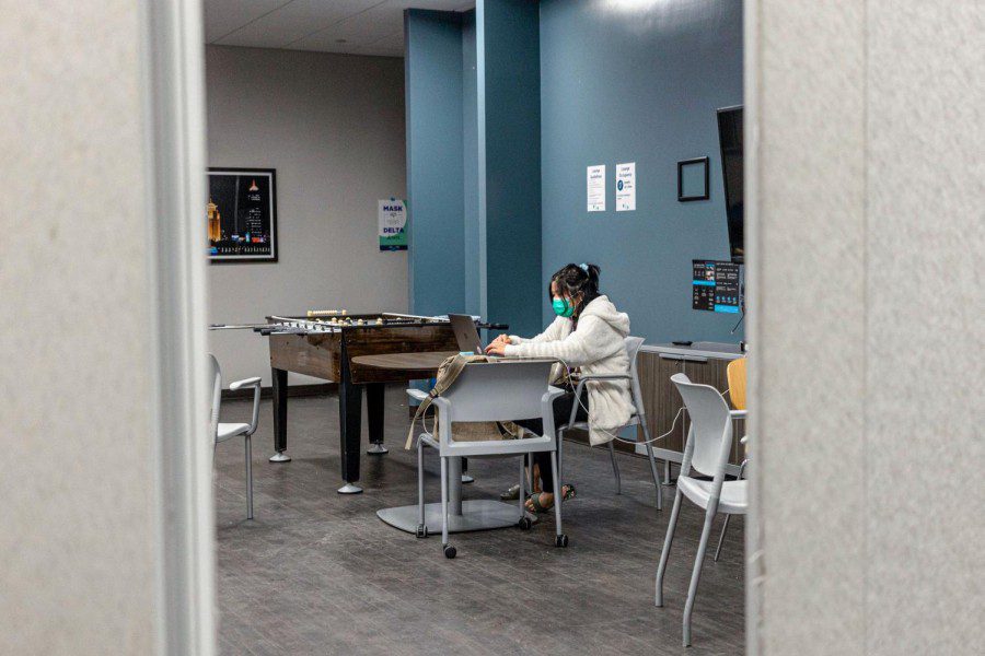 A masked student sits in an NYU common area. She is surrounded by tables, chairs and a foosball table.