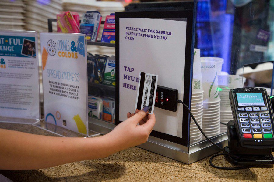 A hand with purple fingernails holds a New York University campus card near a card reader at a dining hall kiosk.
