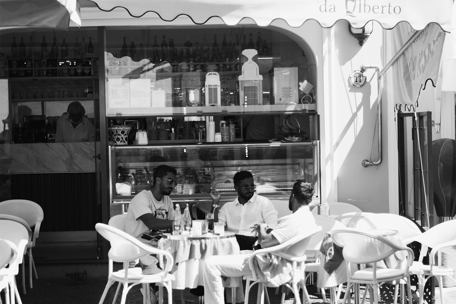 Black-and-white photo of people sitting around a table outside a restaurant in sunshine.