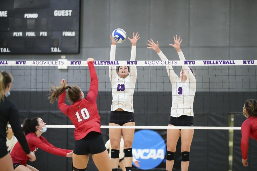 Two members of the women’s volleyball team pass a volleyball ball through the net to the opposing team dressed in red.