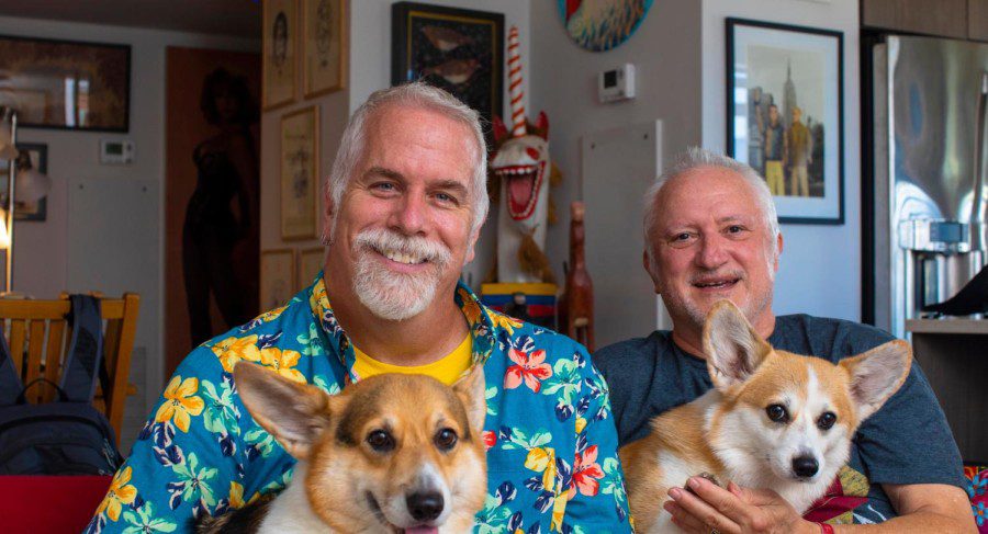 NYU professor John Moran and his husband Kenneth French smile at the camera as they sit on their living room couch with their two corgis. John is wearing a yellow t-shirt under a blue floral dress shirt. Kenneth is wearing a teal-colored t-shirt.