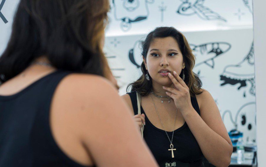 Tori Morales looks into a mirror. They are wearing ear, nose and lip piercings and a necklace with a cross on it.