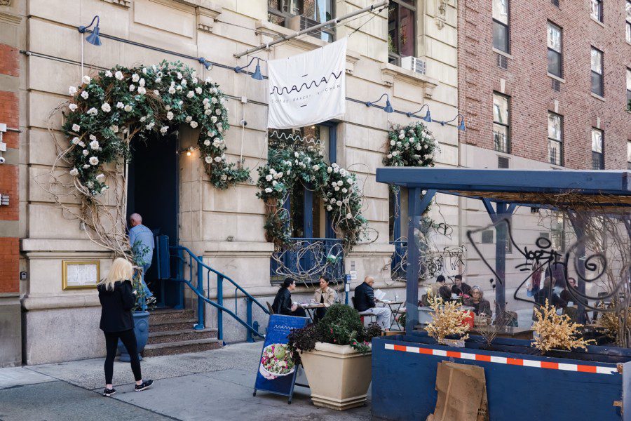 The outdoor dining area on the street outside a cafe with its doors and windows decorated with flowers. A flag with text “MAMAN COFFEE. BAKERY. KITCHEN.” hangs above the entrance.