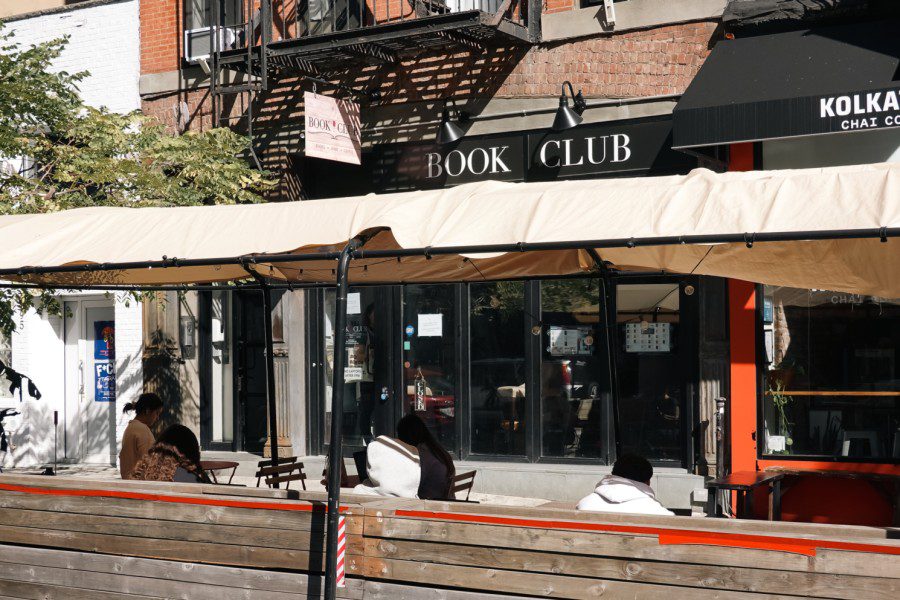 The outdoor dining area on the street outside a cafe with text “BOOK CLUB” written above its entrance with five people sitting outside.