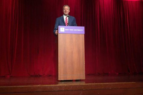Bill de Blasio speaks from behind a podium with a purple banner that reads “New York University” wearing a navy suit with white-and-red striped tie.