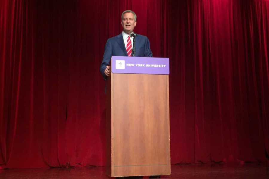 Bill de Blasio speaks from behind a podium with a purple banner that reads “New York University” wearing a navy suit with white-and-red striped tie.
