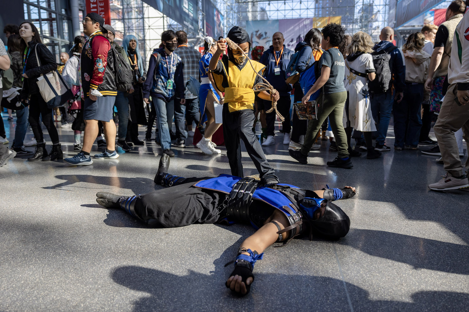 Two cosplayers dressed in Mortal Kombat costumes act out a fight scene. A cosplayer in black-and-blue combat gear lays on the ground. A child in a similar outfit colored black and gold places one foot on the man's stomach.