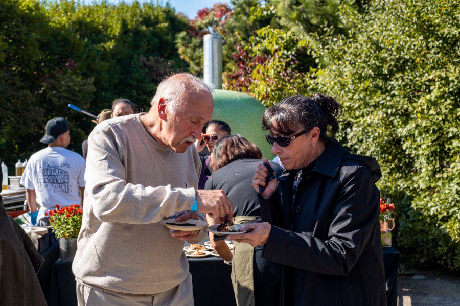 A man in a gray long-sleeved shirt reaches over to a woman's plate. She wears a black coat with sunglasses.