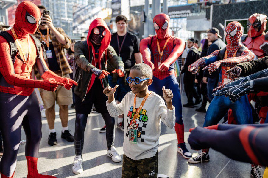 A circle of cosplayers dressed as different versions of Spider-Man surround a child and point at him. From left to right, a person in a red-and-blue classic Spider-Man bodysuit, a person in a black suit with a black-and-red hoodie, another classic Spider-Man, a Spider-Man from the Tobey Maguire movie in a red-and-blue suit with white spider web accents. A child in the center of the circle is dressed in a white shirt that reads Super Mario in rainbow letters, depicting the Mario character in front of a black-and-white checkered rectangle.