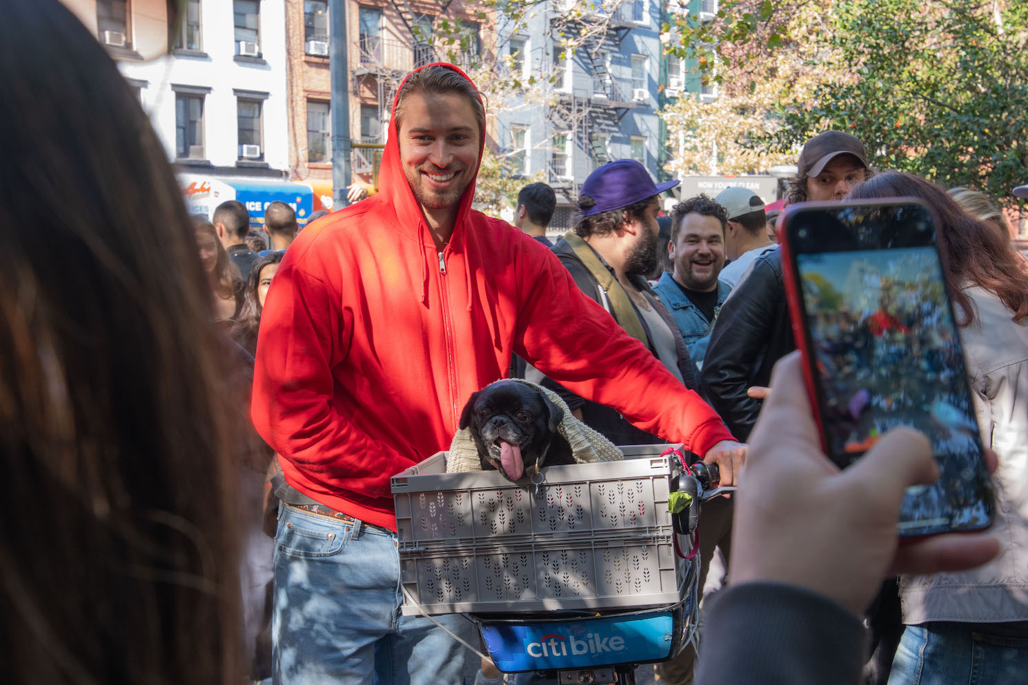 A man wearing a red hooded zip-up sweatshirt smiles for a photo in Tompkins Square Park, holding a Citi Bike that has a gray basket attached to the front. A black pug with its tongue out sits inside the basket. A gray knit blanket is draped over the dog, and there's a hand holding a phone on the right taking a photo of the dog and the owner.