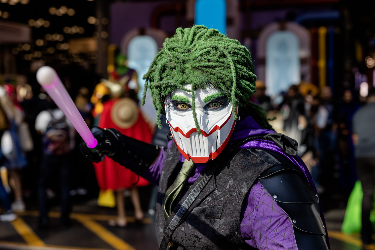 A cosplayer wearing a mechanical mask and green wig holds a purple lightsaber.
