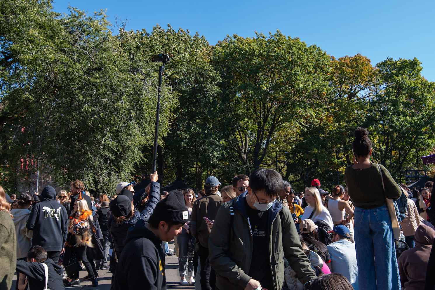A crowd of people gather in the skate park of Tompkins Square Park. A person on the left holds a six-foot-long selfie stick with a phone attached.