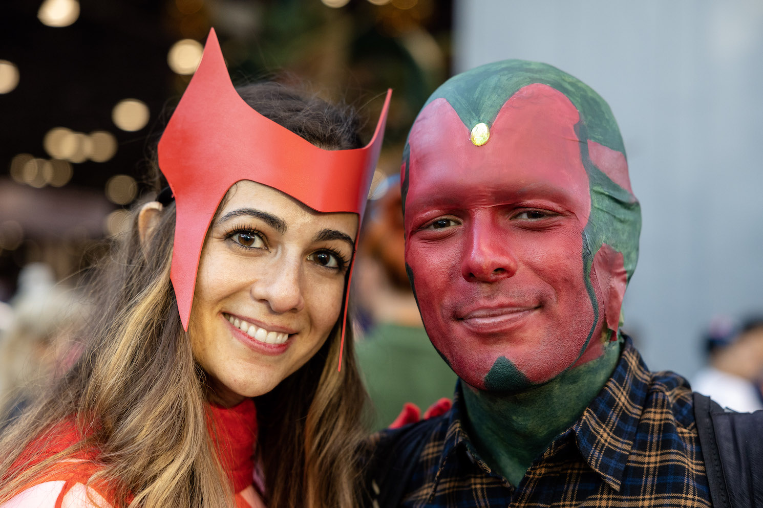 : A couple dressed as the Marvel character “Wanda” and “Vision.” On the left, a woman wears a red crown and matching red turtlenecked suit. On the right, a man with a red-and-green painted face and a yellow gemstone on his forehead wears a flannel shirt.