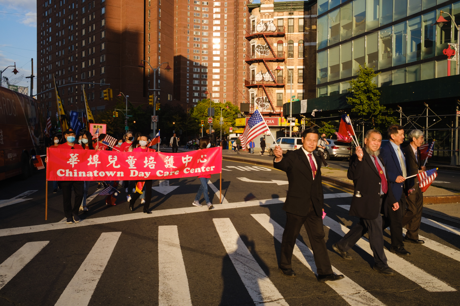 Attendees holding a red banner that reads “Chinatown Day Care Center” marches down Bowery. On the left are four men dressed in black suits holding the American and Taiwanese flags.