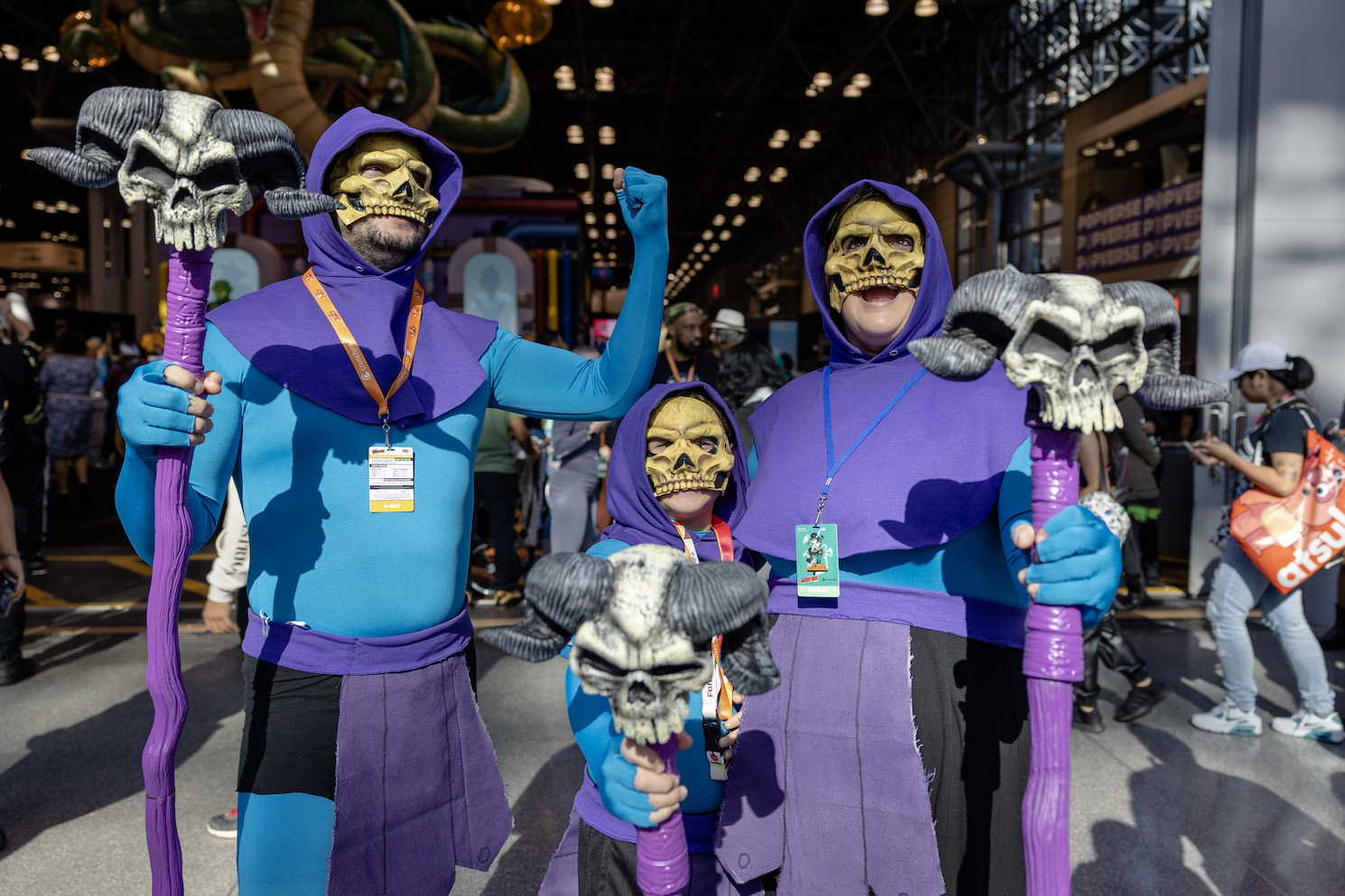 Two adults stand with a child in between them wearing Skeletor costumes. All three wear purple hoods, skeleton masks over the top halves of their faces and blue and purple body suits. They each hold a purple staff with a skeleton head on the top.