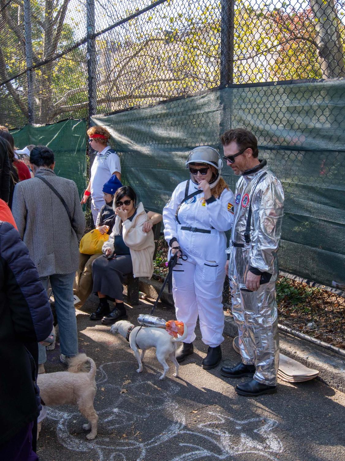 A couple stands with their small, white dog in the Tompkins Square Park skate area. The pair are dressed as astronauts and their dog is dressed as a N.A.S.A. jetpack.